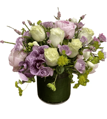 A low, lush floral arrangement in a clear glass vase with a green leaf-wrapped interior, featuring light pink peonies, cream and lavender roses, a large dark purple Phalaenopsis orchid with white stripes, and white freesia, against a white background.