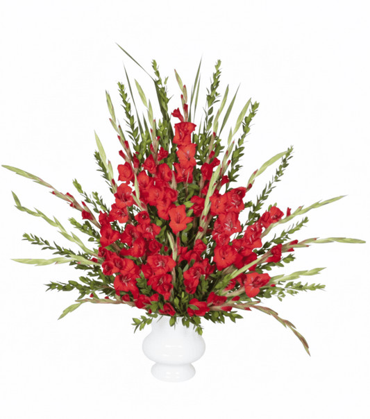 A tall, dramatic floral arrangement in a white pedestal vase, featuring numerous spikes of vibrant red gladiolus flowers and their vertical green leaves, against a pure white background.