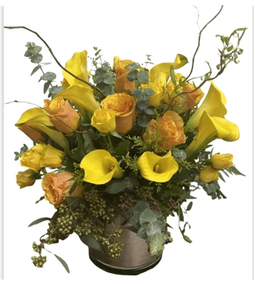 A vibrant floral arrangement in a short, metallic vase, featuring bright yellow calla lilies, peach roses, and green foliage, with tall, curly brown branches for added texture, against a white background.