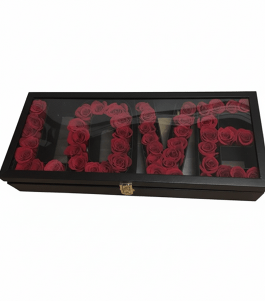 A black rectangular box with a clear lid, containing deep red roses meticulously arranged in separate compartments to spell out the word 'L-O-V-E', with a gold clasp on the front, against a white background.