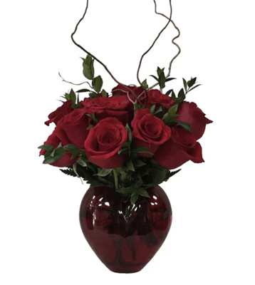 An arrangement of deep red roses and green foliage in a glossy, round-shaped red glass vase, with thin, spiraling curly willow branches accenting the top, against a pure white background.