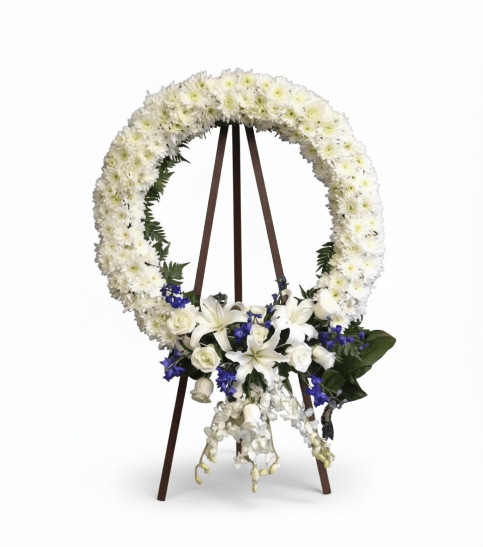 A circular white floral wreath, featuring white chrysanthemums, lilies, and roses, accented with blue delphinium, displayed on a tall wooden easel.
