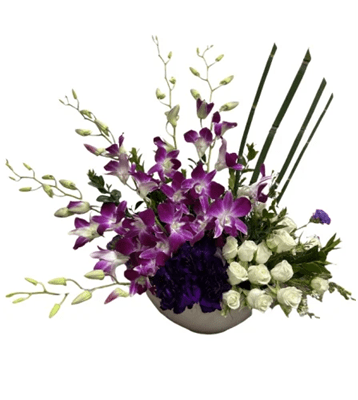 A low, asymmetrical floral arrangement in a white bowl, with a large cascade of purple Dendrobium orchids, a cluster of dark purple carnations, and white spray roses. Tall green stems rise from the right side, against a white background.