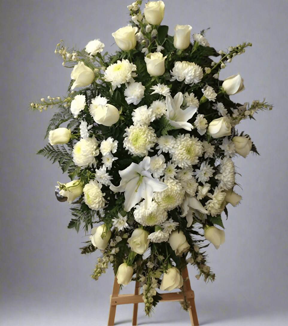 A large, elegant white floral arrangement, named 'Sincere Condolences,' featuring white roses, lilies, and chrysanthemums on a tall wooden easel, set against a plain background. The arrangement is designed for sympathy and remembrance.