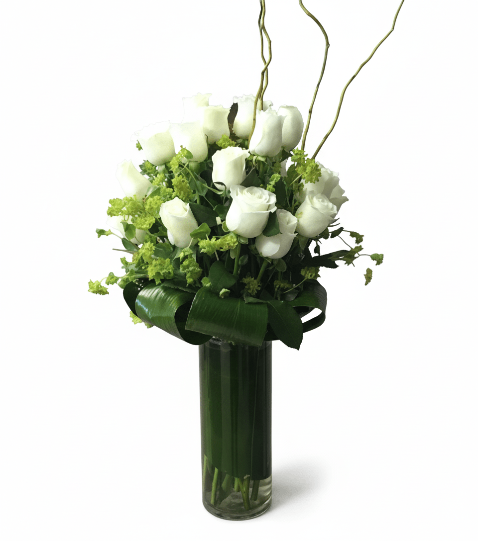 A tall glass vase arrangement featuring elegant white roses accented with delicate green filler flowers, tropical leaves wrapped around the base, and curly willow branches for height and style.