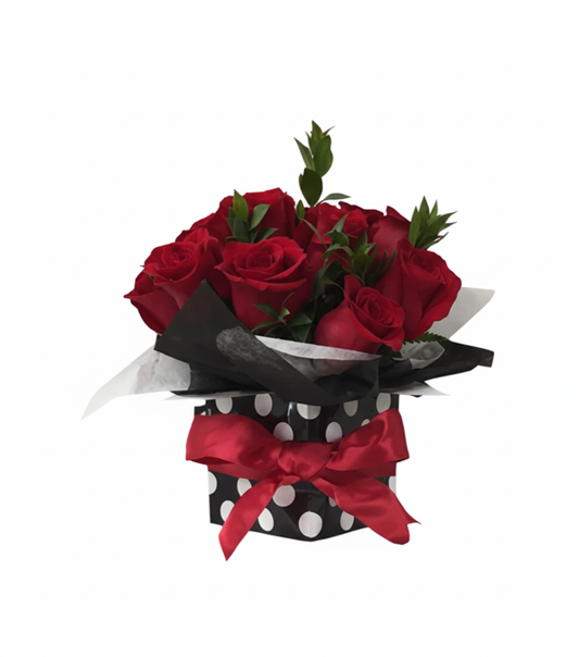 A striking arrangement called "The Royal Roses" featuring a compact bouquet of deep red roses and green foliage, presented in a black box with white polka dots, tied with a bold red satin bow. The background is solid white.