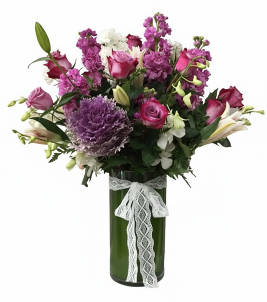 A vibrant and textured floral arrangement named "Lavender Sparkle Jewelry" in a tall, dark green glass cylinder vase adorned with a white lace ribbon, set against a solid white background. The bouquet features a rich mix of purple ornamental cabbage, variegated pink and purple roses, tall spires of purple stock, white lilies (some still in bud), and white dendrobium orchids, with various green foliage.