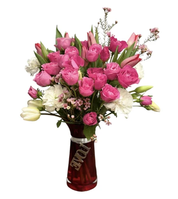 A tall floral bouquet in a reddish-tinted glass vase, featuring pink roses, white tulips, white carnations, and pink filler flowers. A silver wire with the word 'LOVE' is wrapped around the vase, against a pure white background.