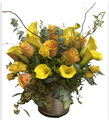 A vibrant floral arrangement in a short, metallic vase, featuring bright yellow calla lilies, peach roses, and green foliage, with tall, curly brown branches for added texture, against a white background.