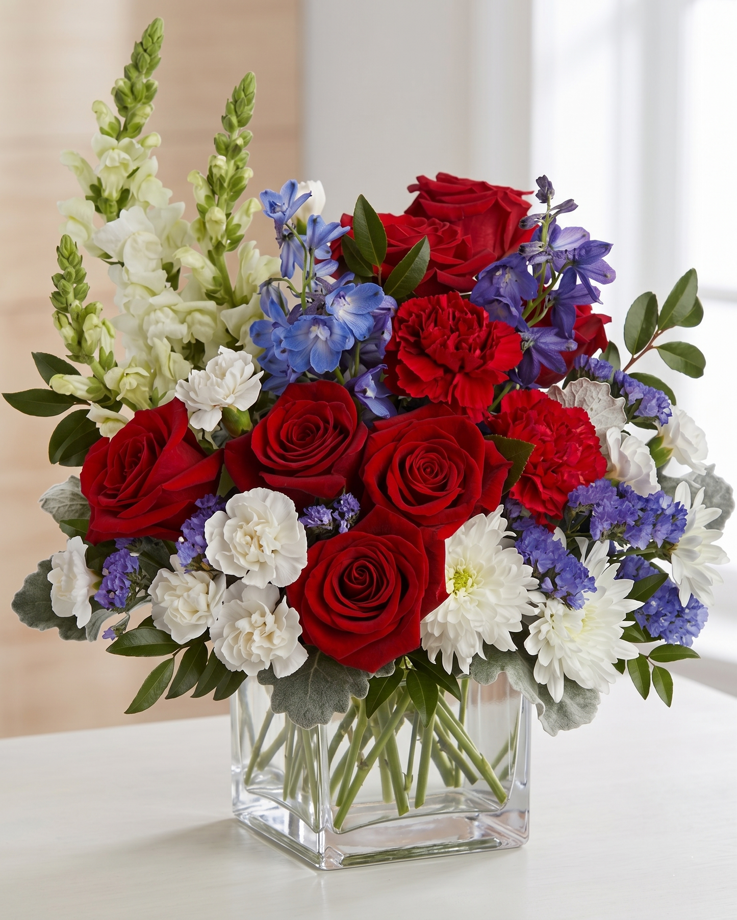 Bouquet of red roses, white flowers, and blue delphinium in a clear glass vase.