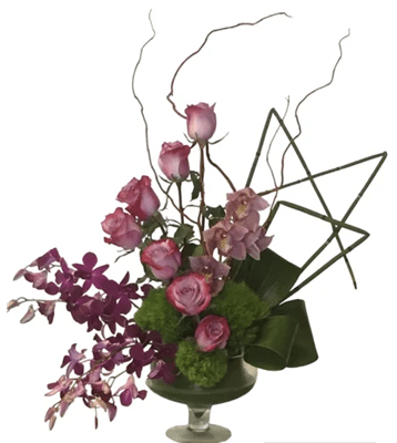 A tall, artistic floral arrangement with pink and lavender roses, light pink and dark purple orchids, accented by green spherical flowers, folded green foliage, and curly willow, in a clear footed glass vase, against a pure white background.
