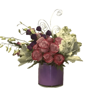 A low, wide floral arrangement featuring pink and purple roses, purple lisianthus, white hydrangea, and green orchid stems, accented with silvery dusty miller foliage and spiraling silver wire, in a purple glass cylinder vase, against a pure white background.