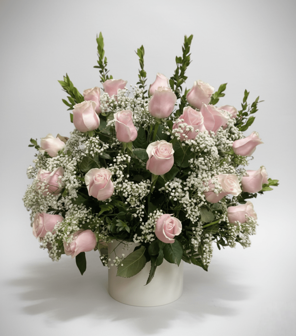 A very full and lush floral arrangement in a short, wide white vase, featuring a dense cluster of light pink roses and a cloud of white baby's breath, against a pure white background.