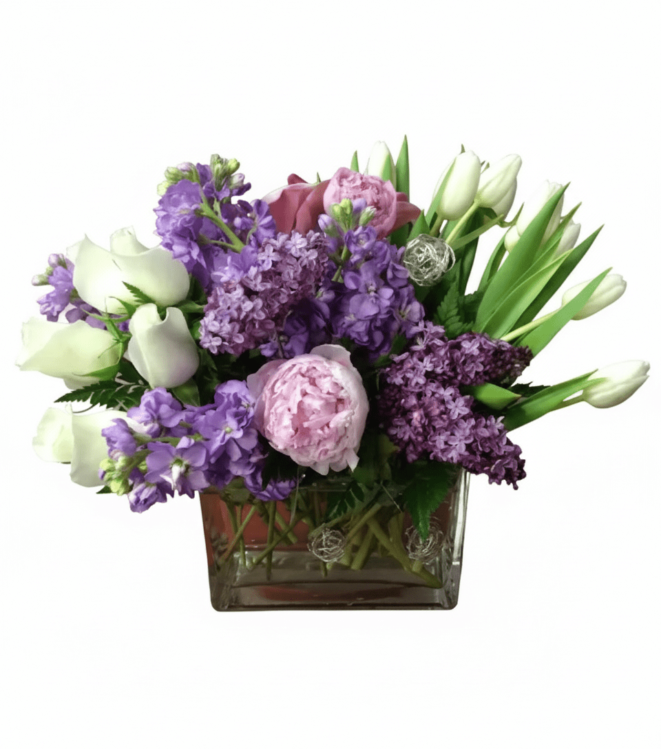 A vibrant floral arrangement featuring purple stock and lilac, pink peonies, and white roses and tulips, presented in a clear glass cube vase.