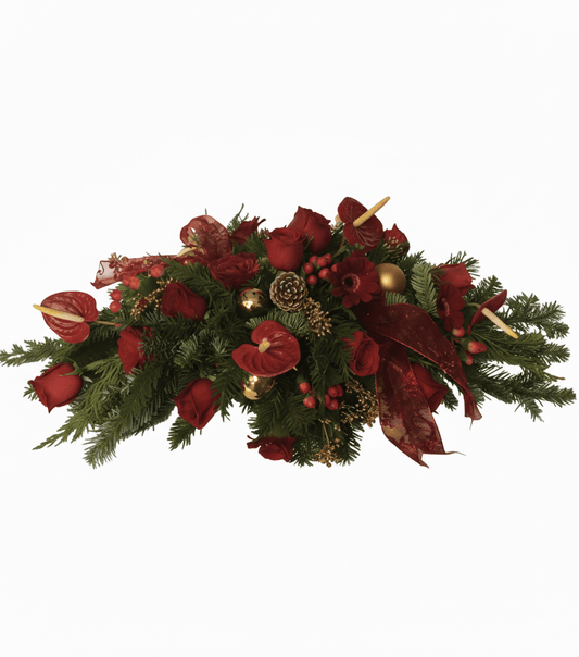 A festive holiday centerpiece featuring lush evergreen branches, red roses, red anthuriums, berries, pinecones, gold ornaments, and decorative red ribbon with candles nestled in the arrangement.