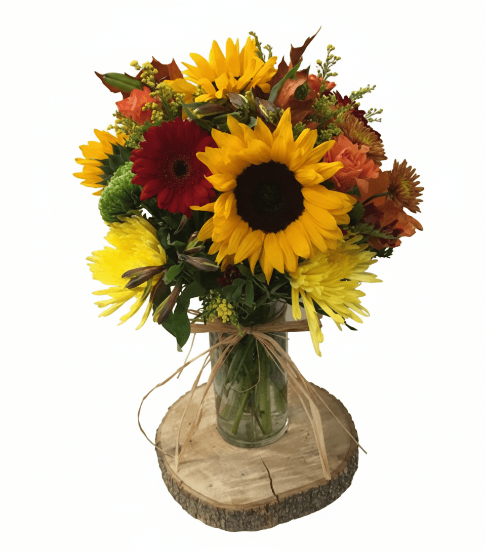 A vibrant autumn-themed floral bouquet named 'Deepest Thanks,' featuring prominent sunflowers, a deep red gerbera daisy, yellow spider chrysanthemums, and various orange and green filler flowers, arranged in a glass jar tied with raffia, all resting on a rustic wooden slice.