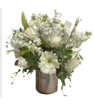 A low, lush floral arrangement in a short silver vase, featuring a prominent white gerbera daisy, white roses, tall white stock flowers, and white calla lilies, complemented by green succulents and button mums, against a white background.