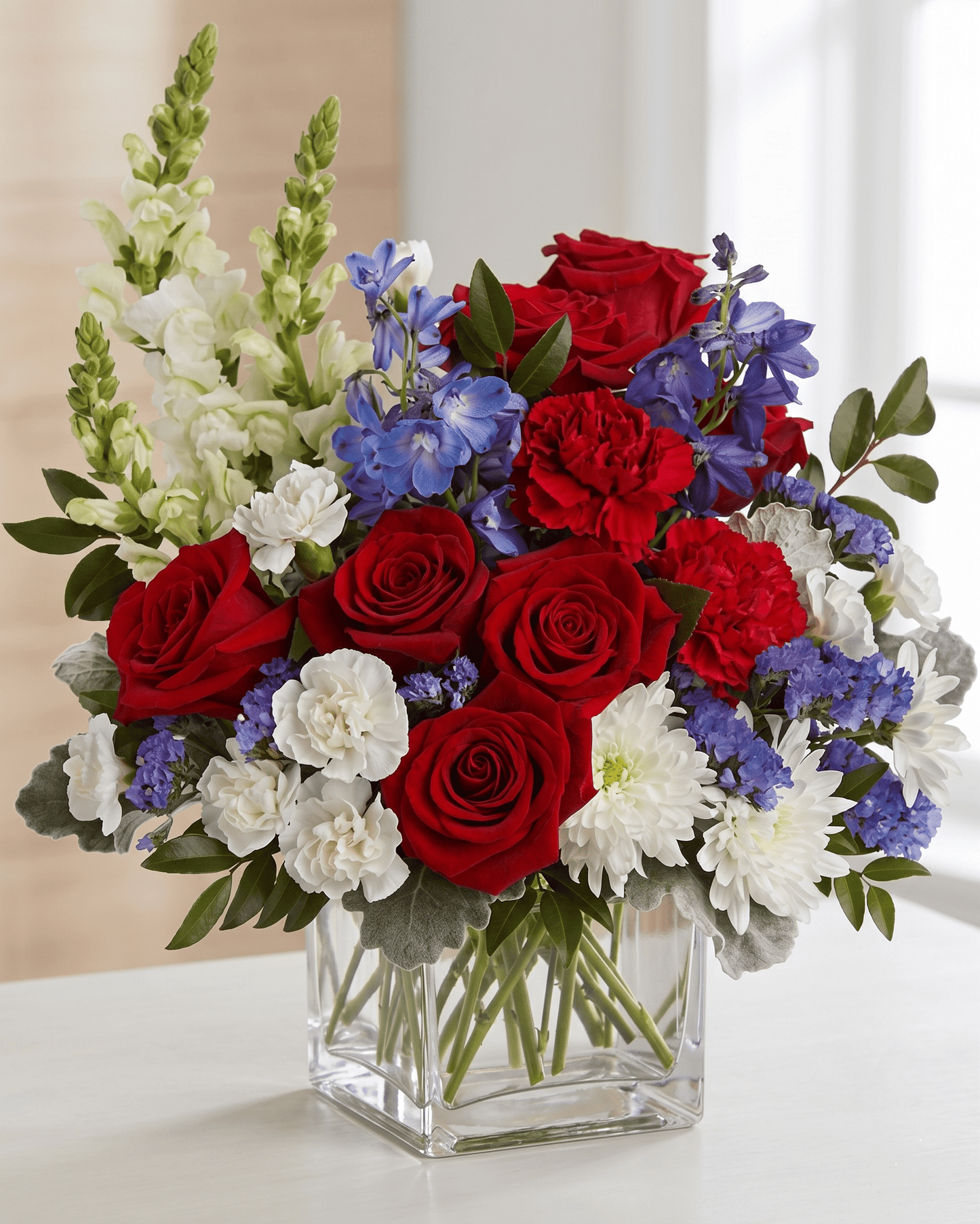 Bouquet of red roses, white flowers, and blue delphinium in a clear glass vase.