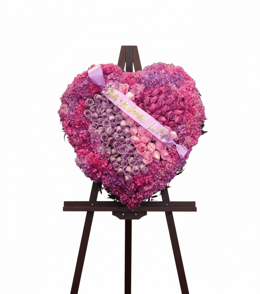 A heart-shaped funeral floral arrangement of pink and purple hydrangeas and roses, with a ribbon that says "Our Beloved Mother," displayed on an easel.