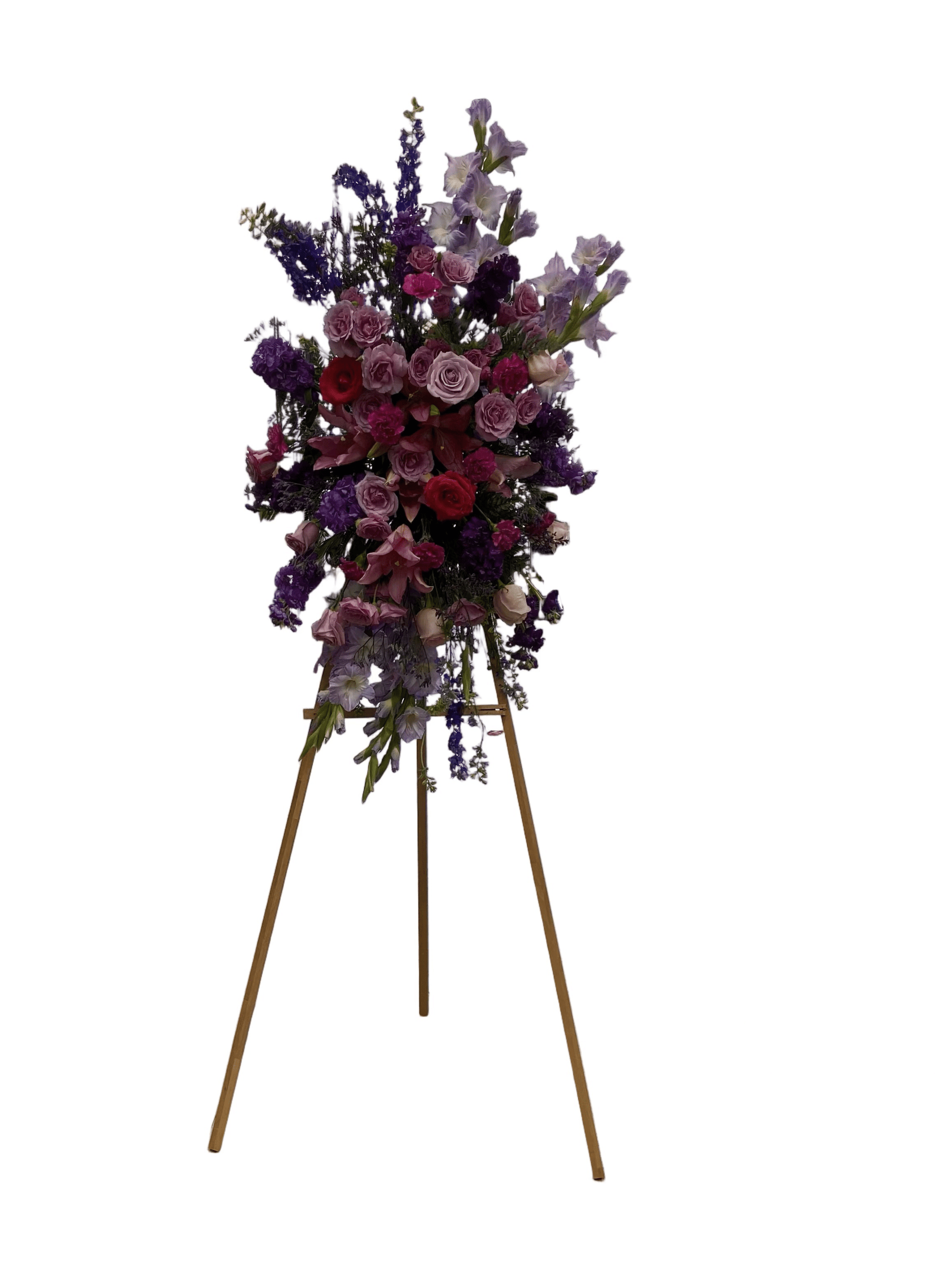 Funeral flower arrangement in shades of purple and pink, presented on a light brown wooden stand, with a clear white background.