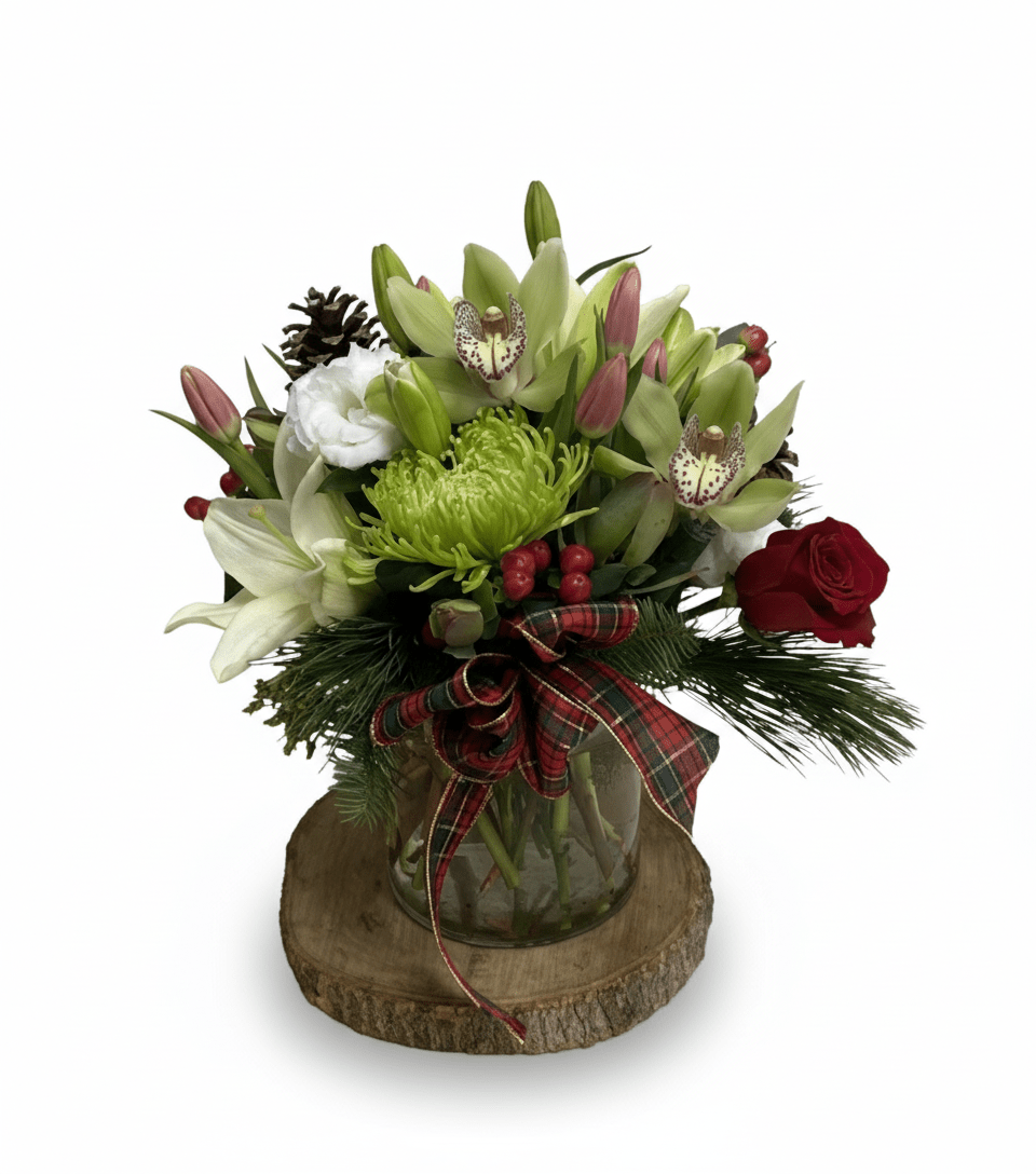 A vibrant holiday floral arrangement featuring green cymbidium orchids, white lilies, pink tulips, a single red rose, green spider mums, pine sprigs, pinecones, and red berries, arranged in a clear glass vase with a red plaid bow, sitting on a wooden slab.