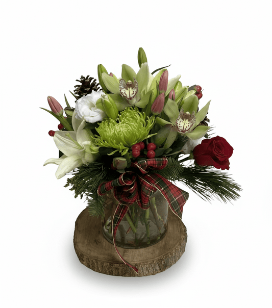 A vibrant holiday floral arrangement featuring green cymbidium orchids, white lilies, pink tulips, a single red rose, green spider mums, pine sprigs, pinecones, and red berries, arranged in a clear glass vase with a red plaid bow, sitting on a wooden slab.