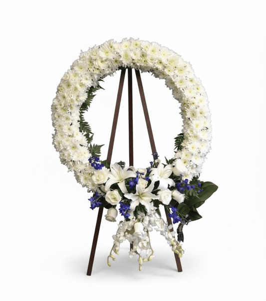A circular white floral wreath, featuring white chrysanthemums, lilies, and roses, accented with blue delphinium, displayed on a tall wooden easel.