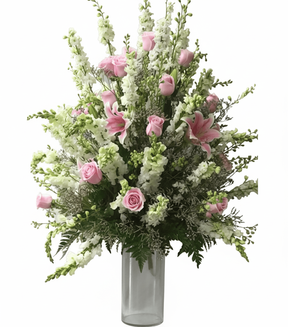 A tall, elegant floral arrangement named "Pink Rose Parade" in a clear glass vase, set against a solid white background. The bouquet features numerous light pink roses and pink stargazer lilies interspersed among towering white delphiniums or snapdragons, with delicate green foliage and filler.