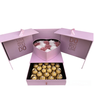A luxurious pink gift box that opens into three sections, with the phrases 'LOVE WHAT YOU DO' and 'DO WHAT YOU LOVE' in metallic text on the side panels. The center section has a top tier with a heart-shaped arrangement of white and light pink roses and a bottom drawer pulled out to reveal a grid of gold-wrapped chocolates, all against a white background.