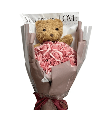 A large, wrapped bouquet with a small, light brown teddy bear at the top, holding a dense cluster of dusty pink rose-shaped soap flowers. The bouquet is wrapped in brown and white paper with the words 'You are my LOVE' printed on it and tied with a maroon ribbon.