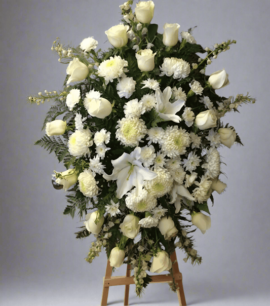 A large, elegant white floral arrangement, named 'Sincere Condolences,' featuring white roses, lilies, and chrysanthemums on a tall wooden easel, set against a plain background. The arrangement is designed for sympathy and remembrance.