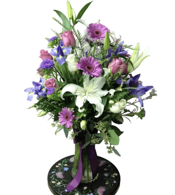 A tall floral arrangement featuring white lilies, purple and blue irises, purple gerbera daisies, and pink roses, with lush greenery and a deep purple ribbon tied around the clear glass vase, set on a decorated stand against a white background.