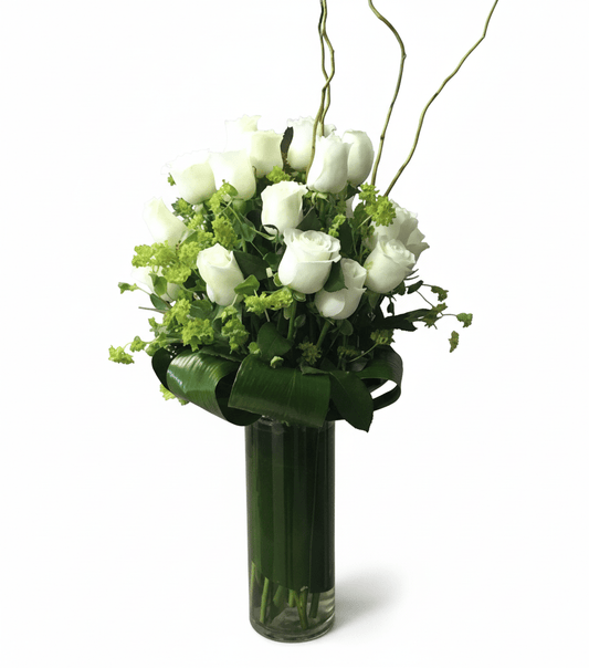 A tall glass vase arrangement featuring elegant white roses accented with delicate green filler flowers, tropical leaves wrapped around the base, and curly willow branches for height and style.