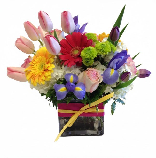 A colorful floral arrangement featuring pink tulips, yellow gerbera daisies, red gerbera daisies, blue irises, green button mums, white hydrangeas, and pink roses in a dark cube vase wrapped with a pink and yellow ribbon.