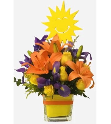 A vibrant floral arrangement featuring orange lilies, purple irises, and yellow roses in a yellow cube vase, with a wooden sun decoration on a stick.
