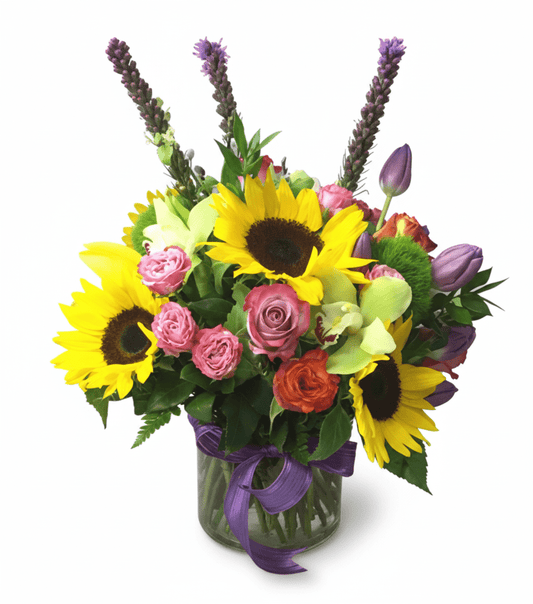 A bright floral arrangement in a clear glass vase with a purple ribbon, featuring bold sunflowers, purple spiky flowers (liatris or lavender), pink and orange roses, green cymbidium orchids, and purple tulips.