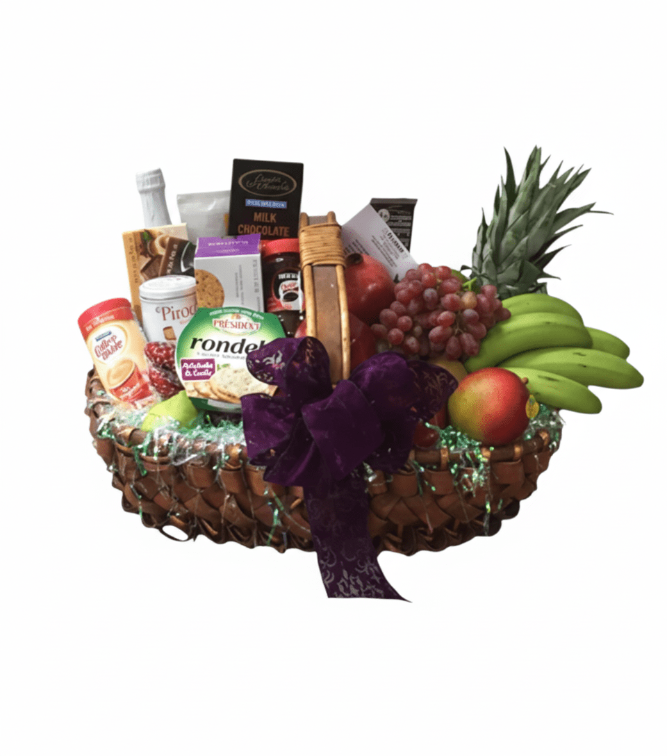 A large, overflowing woven basket against a solid white background, filled with a variety of fresh fruits and gourmet snacks. Visible items include a small pineapple, a bunch of green bananas, red grapes, an apple, a pomegranate, crackers, cheeses (Rondelé), a small bottle of sparkling beverage, various chocolates, and other packaged treats, all adorned with a large purple bow.