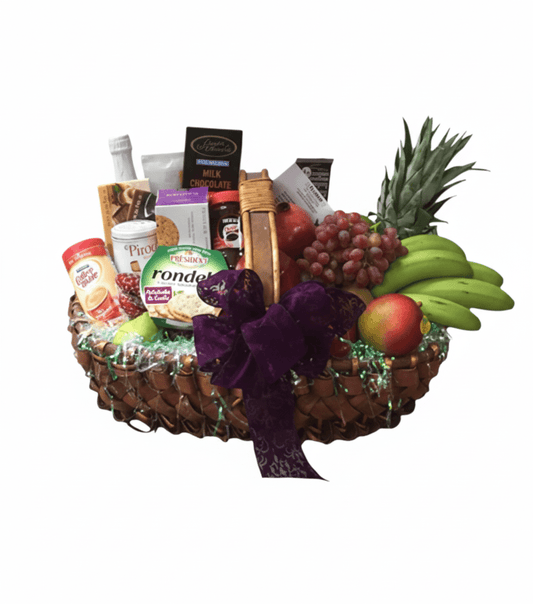 A large, overflowing woven basket against a solid white background, filled with a variety of fresh fruits and gourmet snacks. Visible items include a small pineapple, a bunch of green bananas, red grapes, an apple, a pomegranate, crackers, cheeses (Rondelé), a small bottle of sparkling beverage, various chocolates, and other packaged treats, all adorned with a large purple bow.