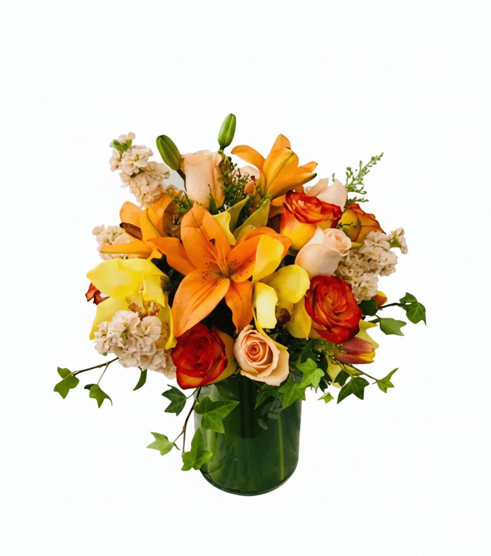A vibrant floral arrangement named 'The Smell Of Orange,' showcasing a rich mix of orange lilies, peach and bi-color orange-red roses, yellow orchids, and light pink stock flowers, with trailing ivy, all presented in a clear glass vase.