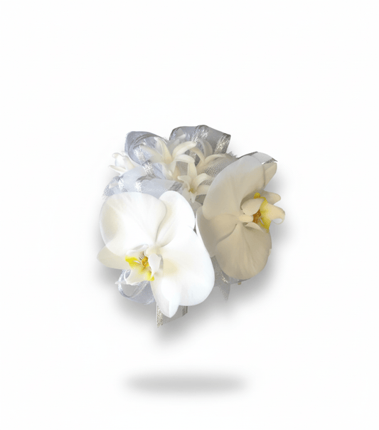 A sophisticated wrist corsage featuring two elegant white Phalaenopsis orchid blooms with yellow centers, accented by delicate white filler flowers and shimmering silver and white sheer ribbons, all against a pure white background.